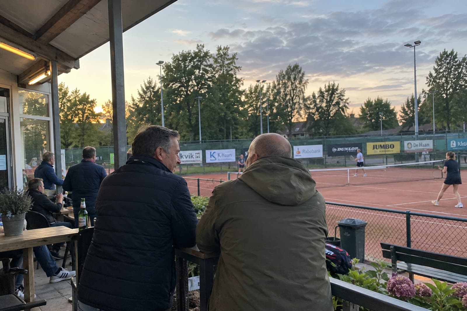 Twee sponsors in gesprek op het terras van een tennisclub, met de banen en de reclameborden op de achtergrond